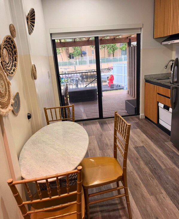 Una cocina con una mesa y sillas de madera, una puerta corrediza de vidrio que da a un espacio exterior con una terraza de madera y una silla roja.