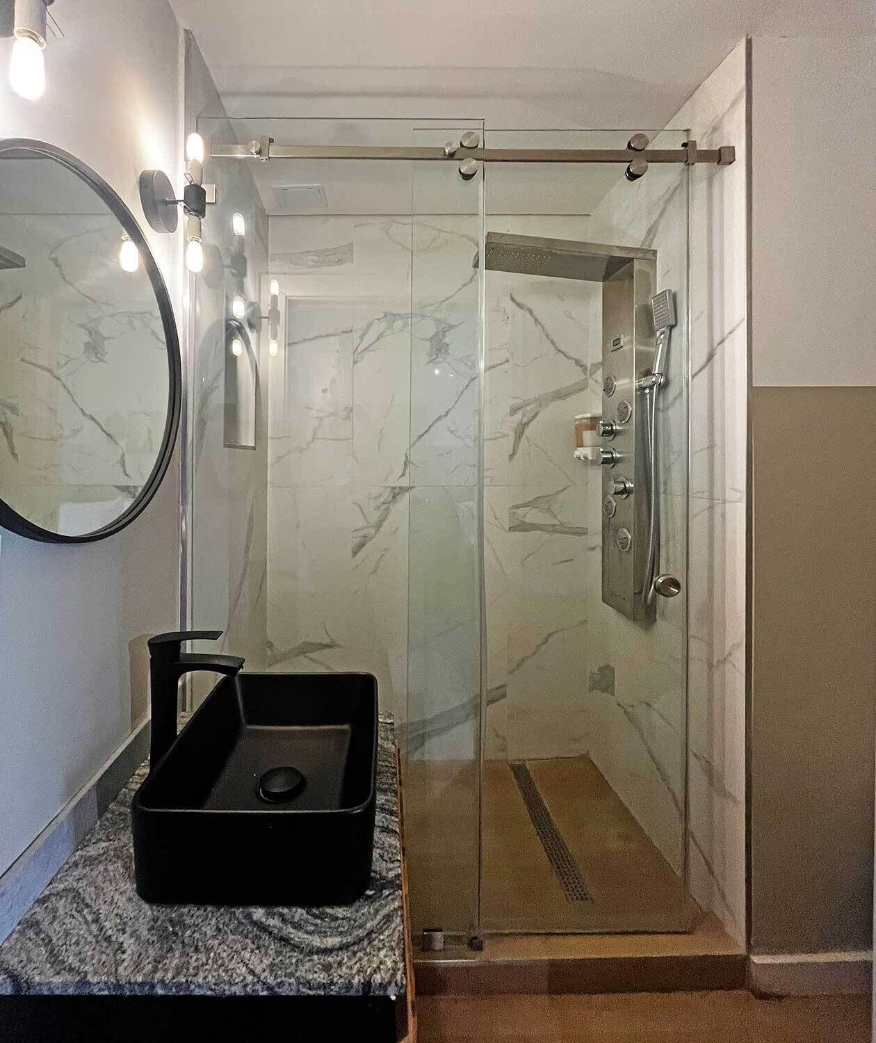 A modern bathroom with a glass shower enclosure, a marble-tiled wall, a round mirror, and a black sink with a tray.