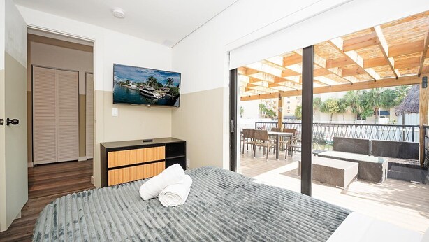 A modern hotel room with a large bed, a flat-screen TV, a wooden dresser, and a balcony with outdoor seating. - Luxury RiverFront by Renzzi (Miami)