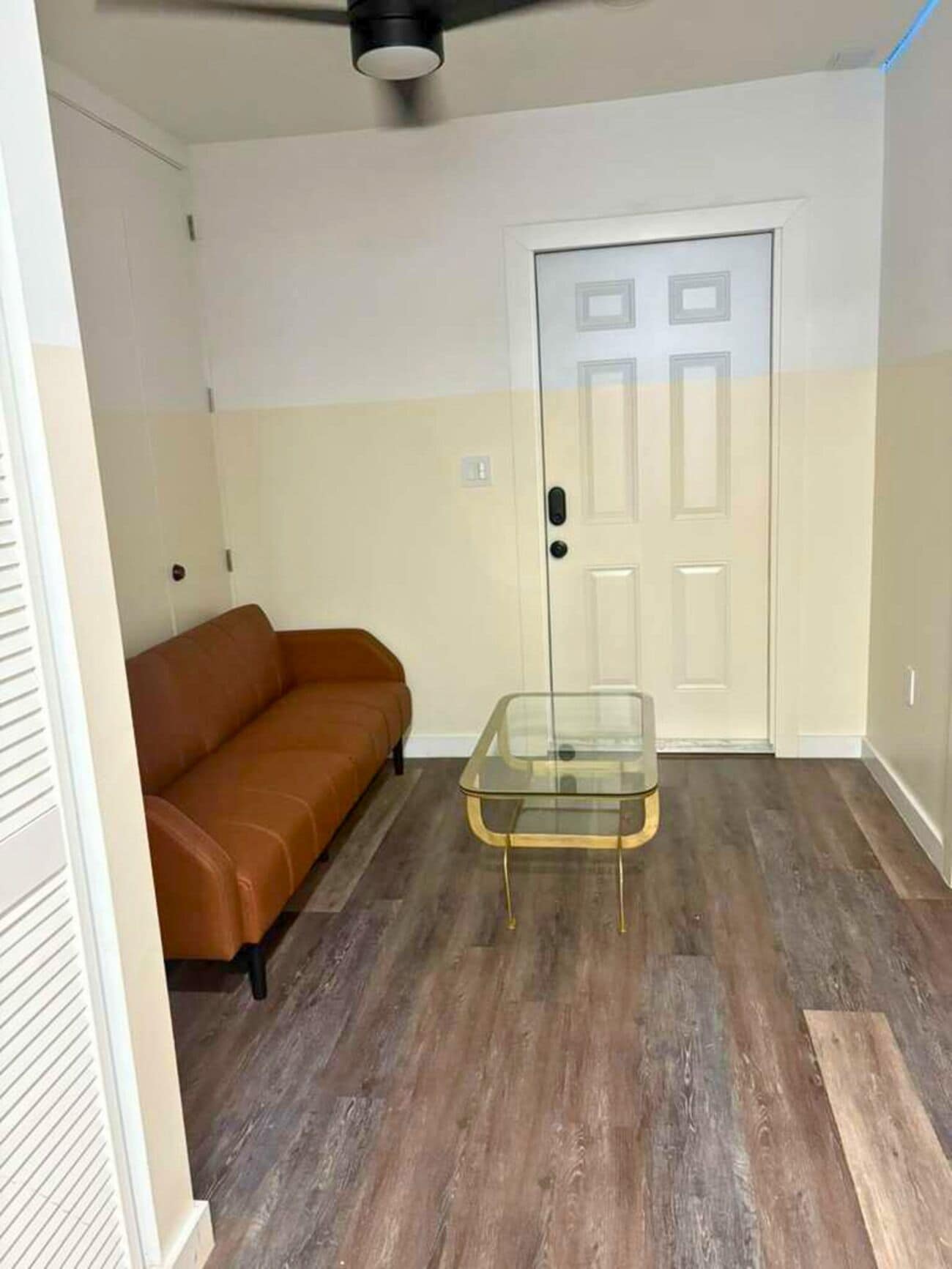 A room with a brown leather sofa, a glass coffee table, a white door, and a ceiling fan.