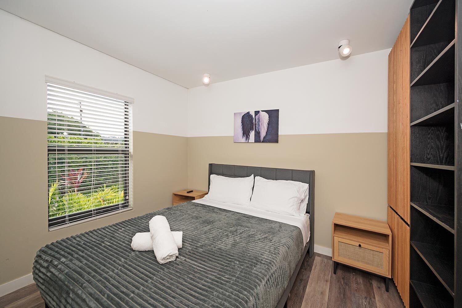 A modern bedroom with a bed, a nightstand, a window with blinds, a wall-mounted shelf, and a ceiling light.