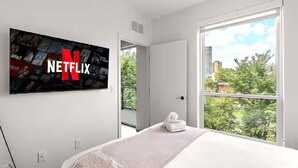A modern hotel room with a large wall-mounted TV displaying the Netflix logo, a bed with a white blanket, and a view of greenery through a large window.