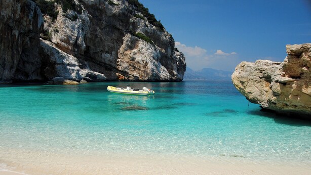 Een strand met helder turquoise water, een klein bootje en rotsachtige kliffen.