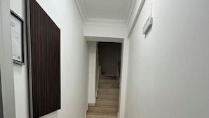 A narrow hallway with a wooden door on the left, a yellow trash bin, and a staircase leading to an upper floor. - İSKELE APART AVŞA  (Marmara)