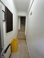 Ein schmaler Korridor mit einer Holztür links, einem gelben Mülleimer und einer Treppe, die zu einer oberen Etage führt.