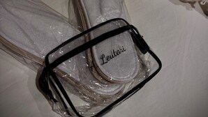 Un par de zapatillas blancas con una tira negra y una etiqueta de nombre, sobre una superficie blanca junto a una toalla enrollada.