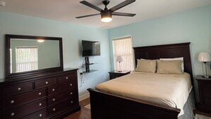 Een slaapkamer met een bed, een dressoir, een plafondventilator, een televisie en een raam met jaloezieën.