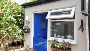 A small house with a blue door and window, white exterior walls, and a small garden area with plants and flowers. - Kenny Cottage (Dawlish)