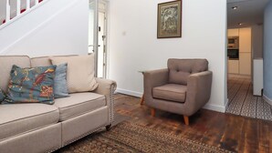 A living room with a beige sofa, patterned cushions, a brown armchair, a wooden floor, a patterned rug, and a staircase with white railings. - Kenny Cottage (Dawlish)