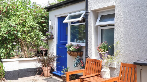 A small, white house with a blue door and windows. There are potted plants and a wooden bench outside. - Kenny Cottage (Dawlish)
