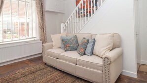 A living room with a beige sofa, patterned cushions, a wooden floor, a white staircase, and a window with curtains. - Kenny Cottage (Dawlish)
