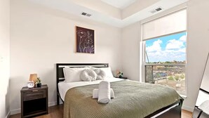 A modern bedroom with a large bed, a nightstand, a framed floral artwork, and a window with a city view. - Stylish 1B in DT Minneapolis, Minutes to Minneapolis Convention Center & US Bank (Minneapolis)
