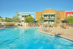 En swimmingpool med liggestole og parasoller, omgivet af et hegn og bygninger med altaner.