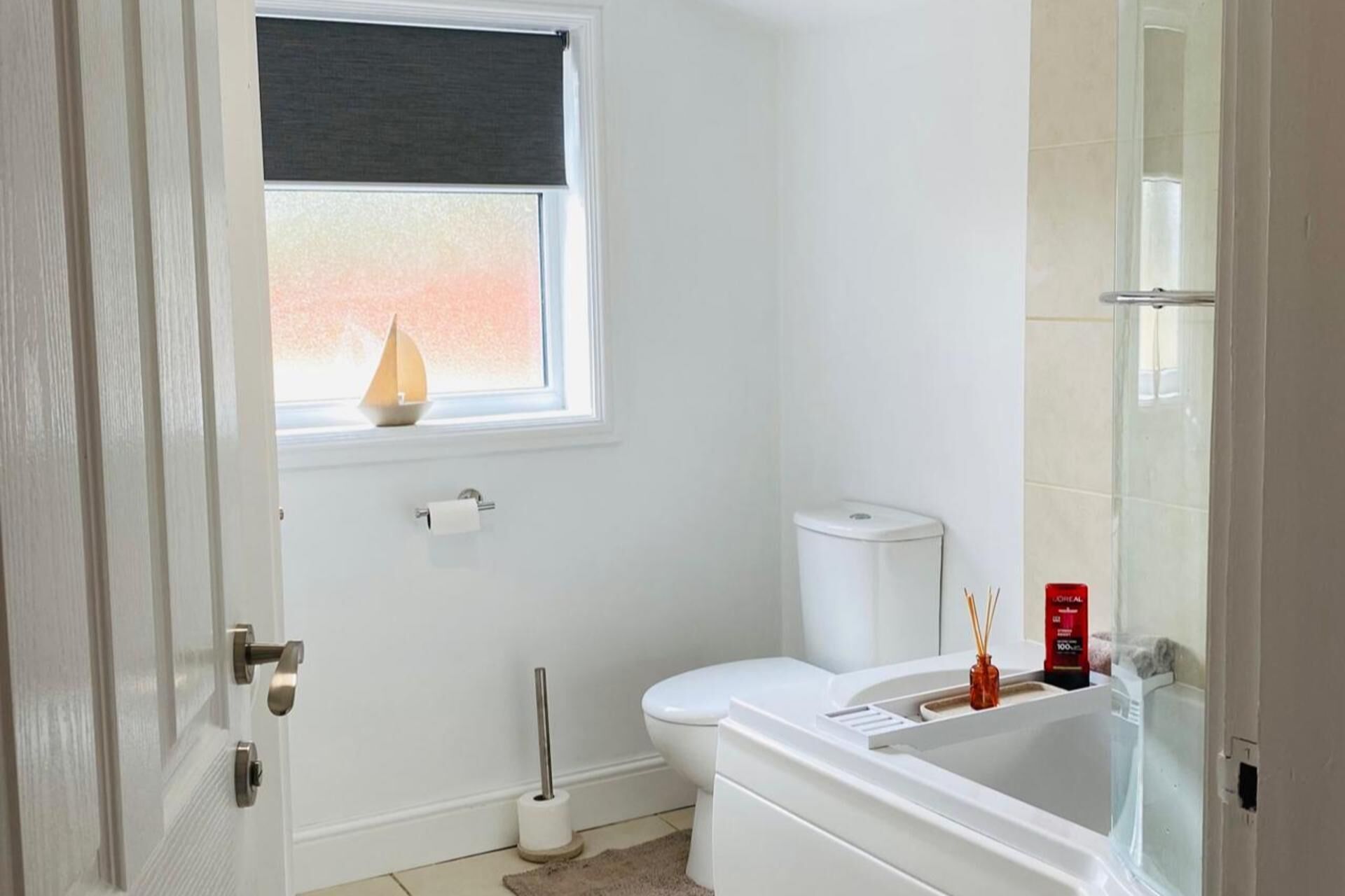 A bathroom with a toilet, a sink, a bathtub, a window with a sailboat decoration, and a toilet paper holder.