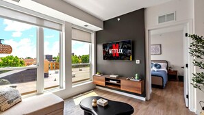 Una sala de estar moderna con un televisor grande montado en la pared mostrando el logotipo de Netflix, un sofá, una mesa de centro y una cama con cabecera azul.
