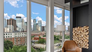 Una sala moderna con ventanas amplias que ofrecen vista a la ciudad, un sillón de cuero, una lámpara de pie y un perchero de pared para leña.