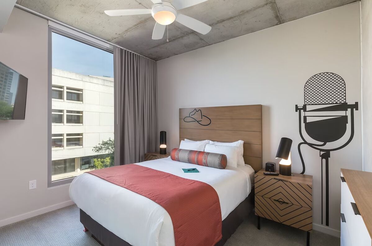 Une chambre d’hôtel avec un grand lit, un ventilateur de plafond, une table de chevet avec une lampe et une vue sur un bâtiment à l’extérieur.