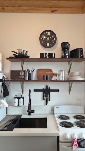 A kitchen with a clock, stove, shelves with kitchenware, and a coffee maker. - Cozy 1BR Tiny Home near Downtown & NB Rivers (New Braunfels)