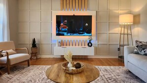 Una sala de estar moderna con un televisor de pantalla plana montado en la pared, una mesa de centro redonda de madera con objetos decorativos, un sofá blanco y una lámpara de pie.