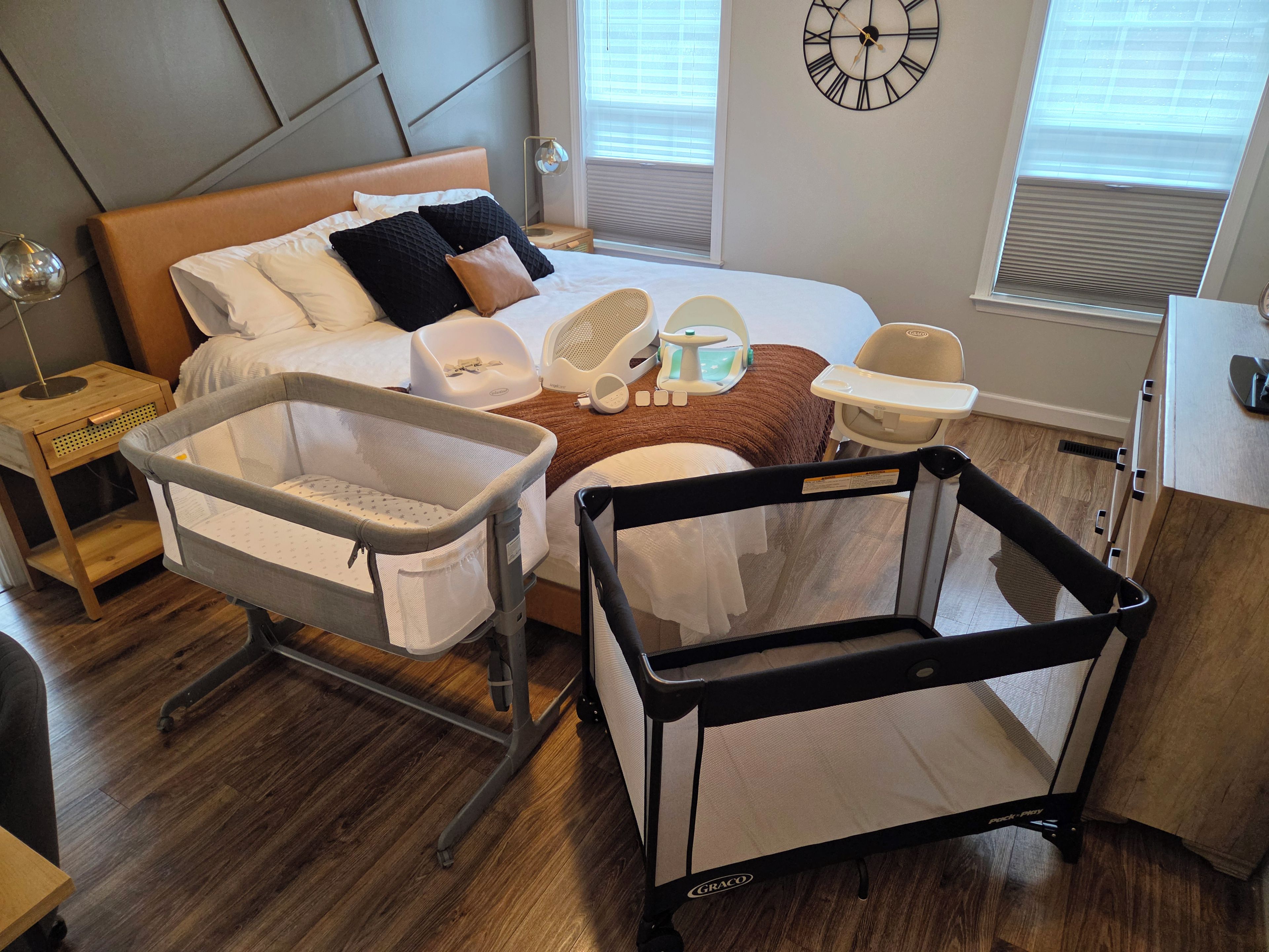 A hotel room with a bed, a changing table, a baby rocker, a clock, and a window with blinds.