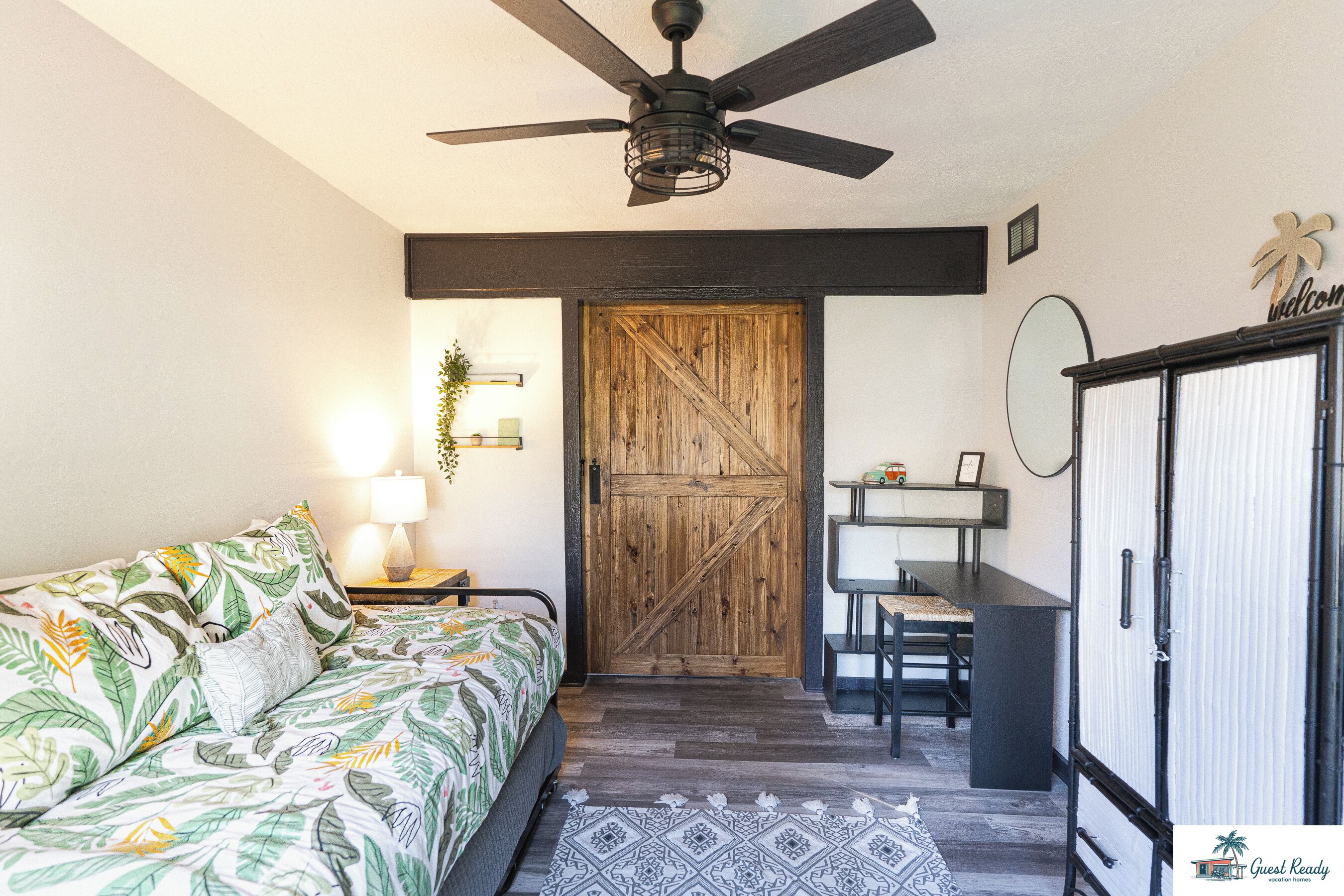 Un dormitorio con cama, escritorio, armario, ventilador de techo y puerta de madera.