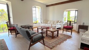A spacious living room with a sofa, chairs, a coffee table, and a dining table. There are large windows providing natural light and a view of greenery outside. - L'appartement Montecristo (Aléria)