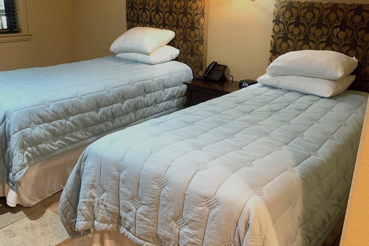 Ein Hotelzimmer mit zwei Betten, jeweils mit weiß gestopfter Bettdecke und Kissen. Zwei Wandlampen und zwei gerahmte Blumenbilder hängen über den Betten.