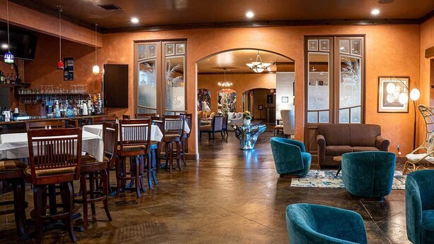 Un espace bar avec des chaises et des tables en bois, un comptoir de bar orné de différentes bouteilles, et un coin salon avec des chaises bleues et un canapé marron.