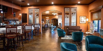 Una zona de bar con sillas y mesas de madera, una barra con diferentes botellas y un área de estar con sillas azules y un sofá marrón.
