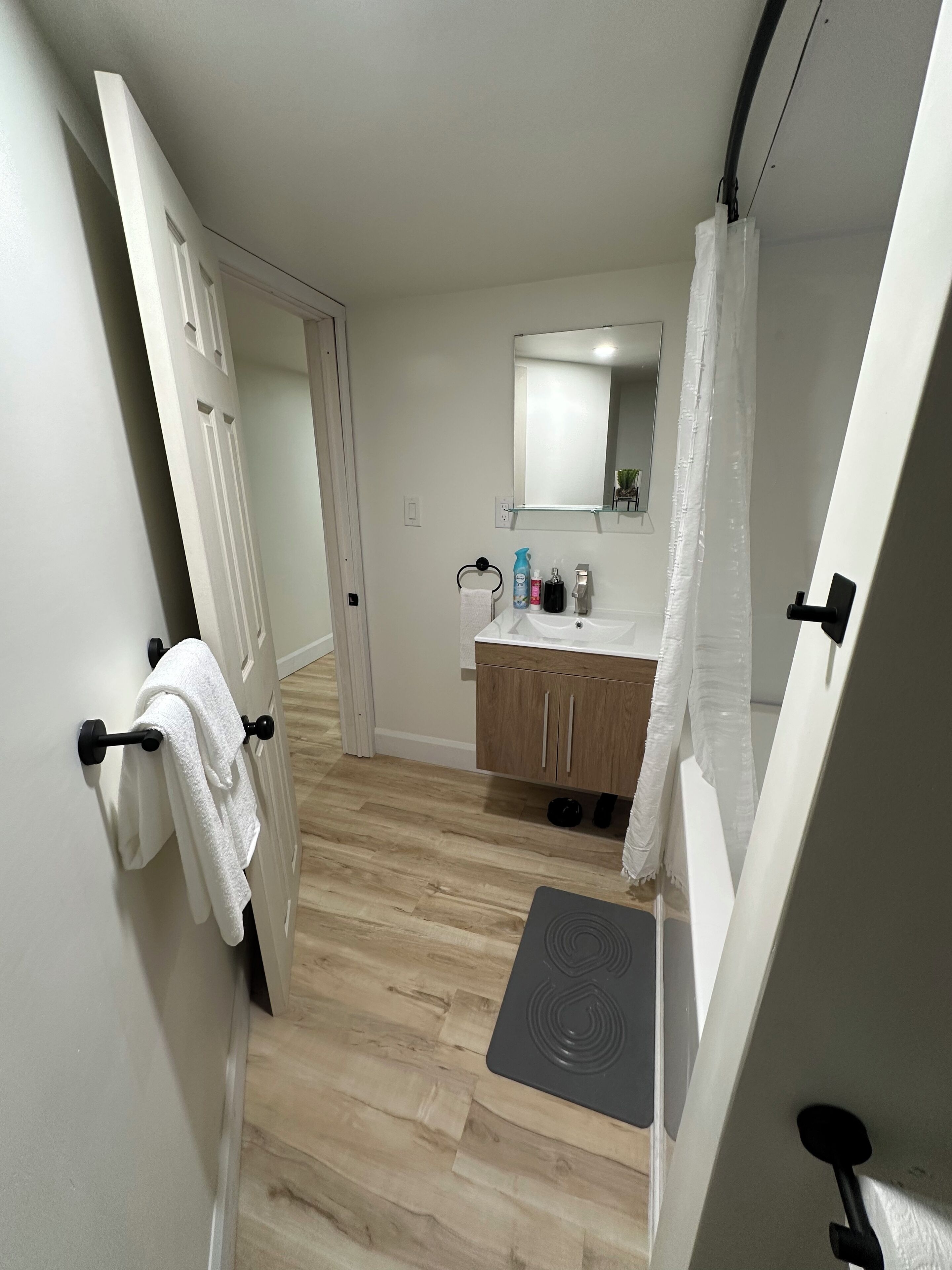 Ein schmaler Flur mit Holzboden, der zu einem Badezimmer mit Waschbecken, Spiegel und Handtuchhalter führt.