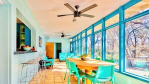 Un coin repas avec une table en bois et des chaises vertes, de grandes fenêtres donnant sur la verdure et un ventilateur de plafond.