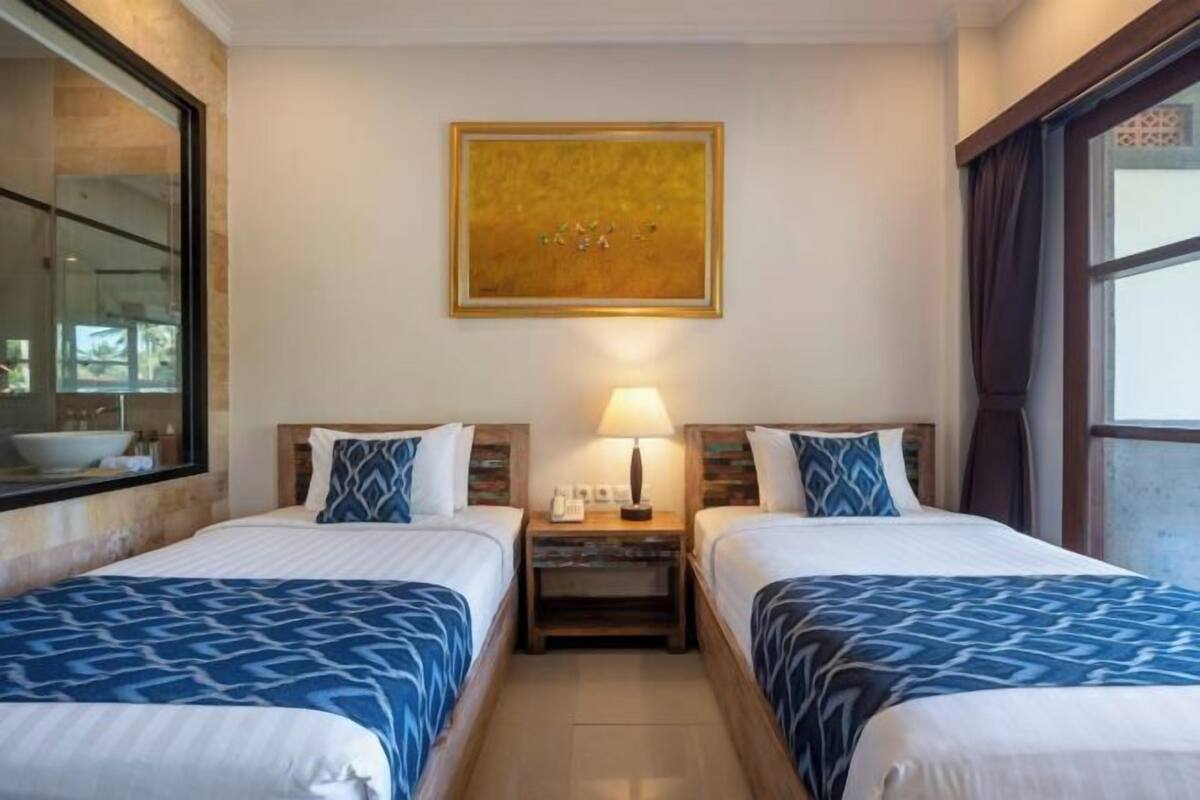 Une chambre d’hôtel avec deux lits, une table de chevet avec une lampe, un tableau au mur et une grande fenêtre.