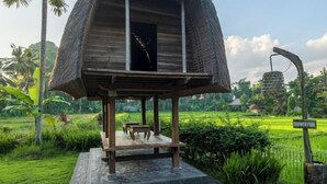 Eine Holzhütte mit einem Reetdach, einem kleinen Tisch und einer Bank auf einer Steinterrasse, umgeben von üppigem Grün und einem Schild, das einen Duschteich anzeigt.