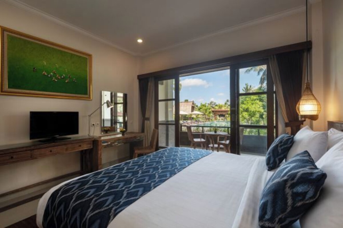 A hotel room with a bed, a desk with a television, a large window, and a balcony with outdoor furniture.