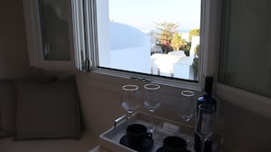 A window with a view of a white building and a tree. There are two wine glasses and a bottle on a tray. - Santozen Suites (Santorini)