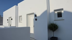 A modern white building with a flat roof, a small tree in a planter, and a door with a number plate. - Santozen Suites (Santorini)