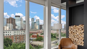 A modern living room with large windows offering a city view, a leather armchair, a floor lamp, and a wall-mounted log display. - Luxury 2Bd Condo DT Minneapolis, Minutes From Convention Center & US Bank Arena (Minneapolis)
