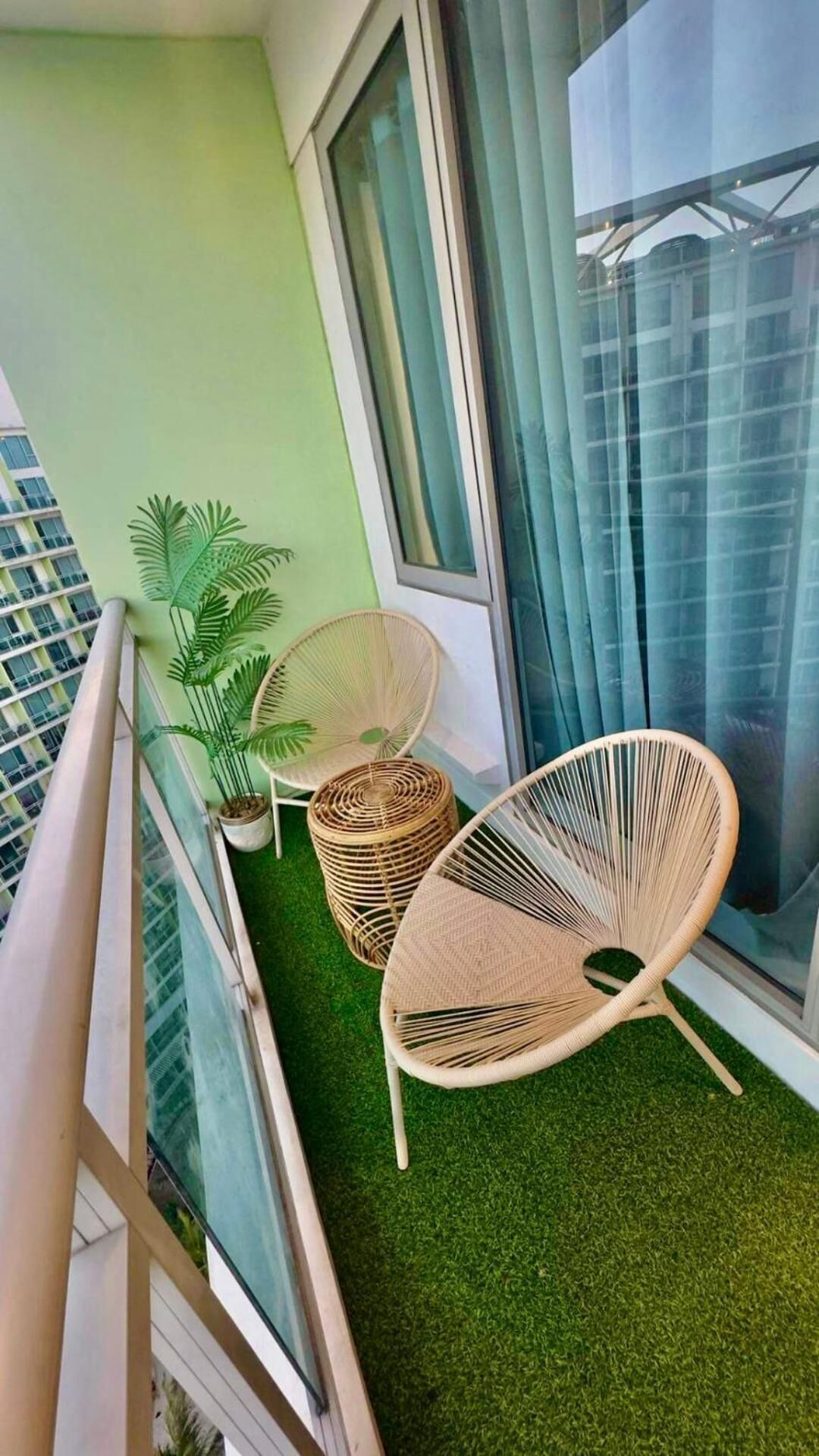A balcony with artificial grass, two wicker chairs, a potted plant, and a view of a cityscape.