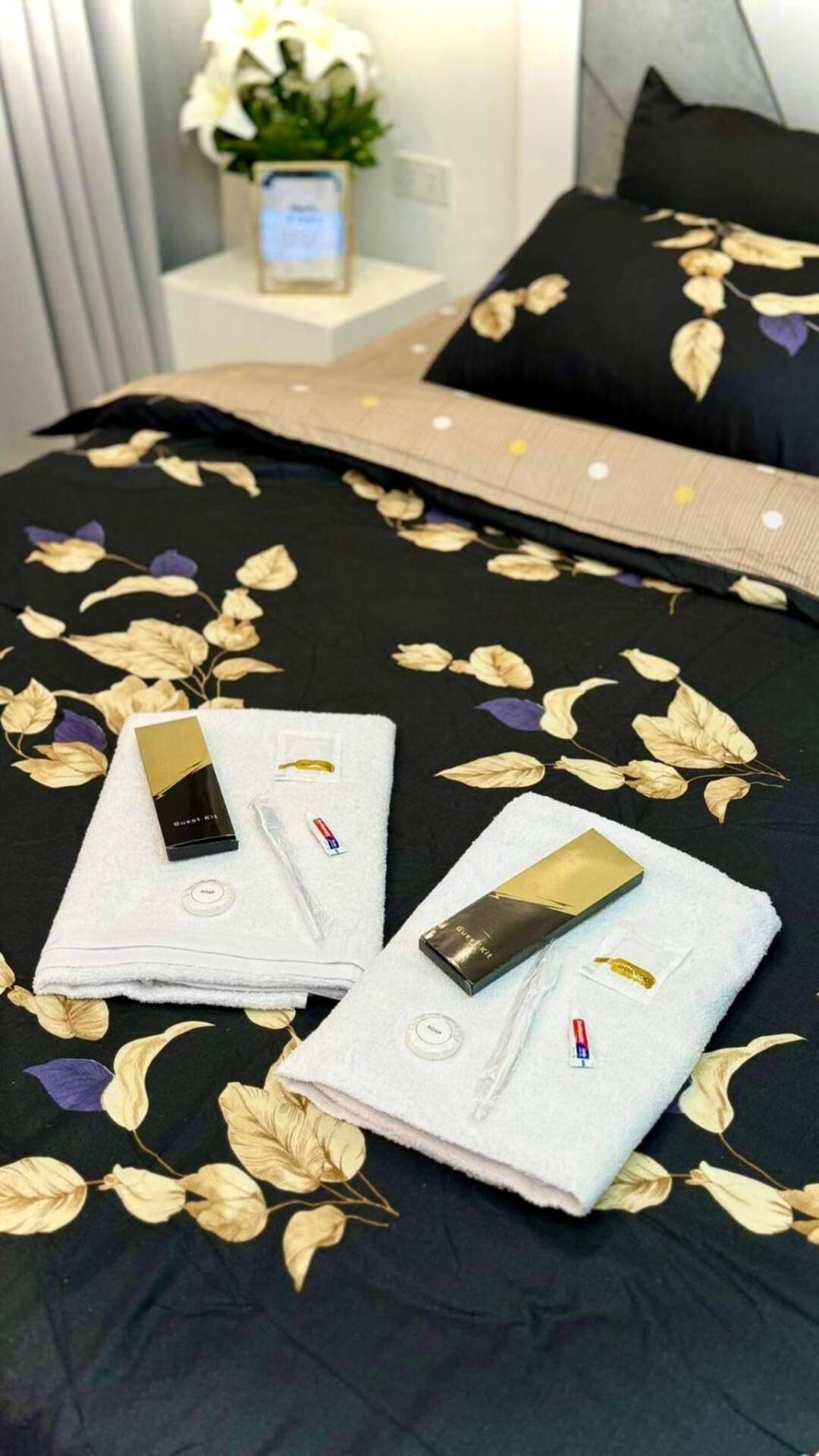 A neatly made bed with a floral bedspread, two towels, and a guest kit on a white pillow.