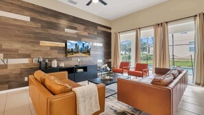 A modern living room with a sofa, armchairs, a coffee table, and a TV mounted on a wooden wall. Large windows offer a view of the outdoors. - Disney Retreat w/ Wizard & Galaxy Rooms (Kissimmee)