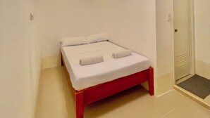 Quarto pequeno com uma cama de solteiro, estrutura vermelha, roupa de cama branca e porta com olho mágico.