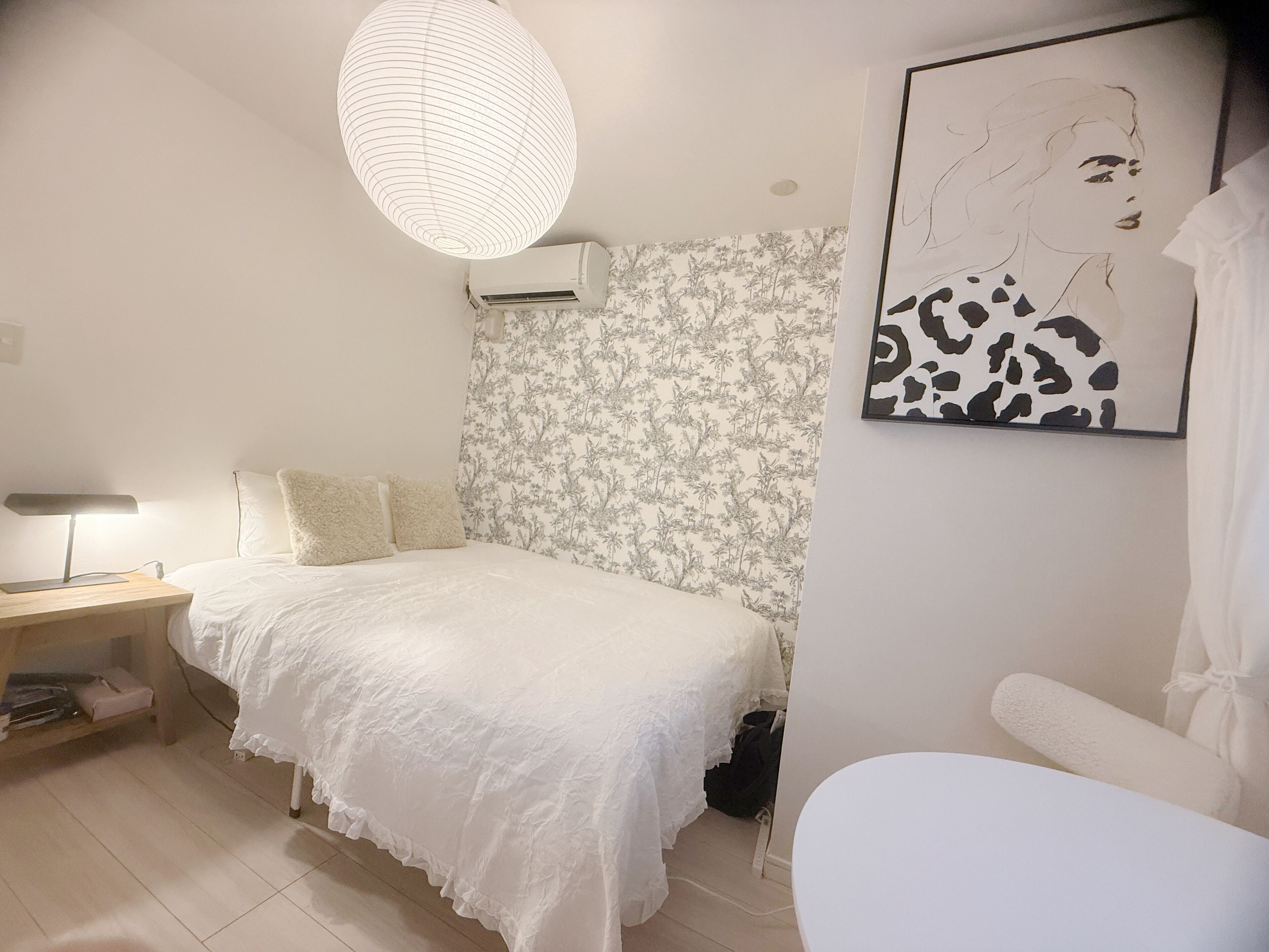 A bedroom with a bed, a bedside table with a lamp, a wall-mounted air conditioner, and a framed artwork on the wall.