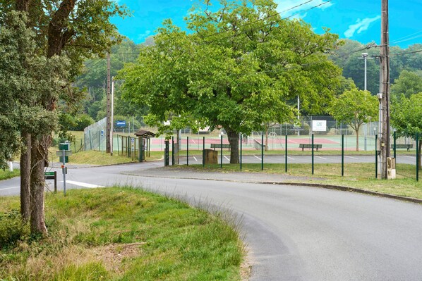 A curved road with a tree-lined path, a fenced sports court, and a bench.