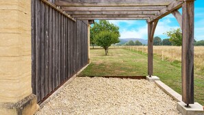 Un espacio al aire libre cubierto, con suelo de grava, vigas de madera y vista a un árbol y un campo cubierto de hierba.