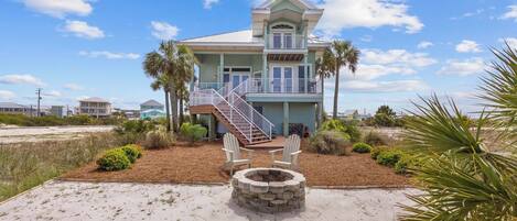 A two-story beachfront house with a stone fire pit, a staircase leading to a balcony, and a view of the beach.