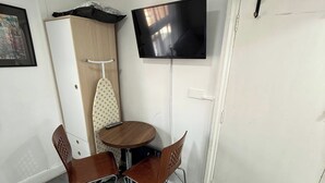 A small room with a wooden cabinet, an ironing board, a round table with two chairs, and a mounted flat-screen TV.