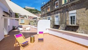 Une terrasse sur le toit, agrémentée de deux chaises longues colorées, d’une petite table et d’une poubelle. La terrasse est bordée d’un mur blanc et d’un bâtiment en pierre.