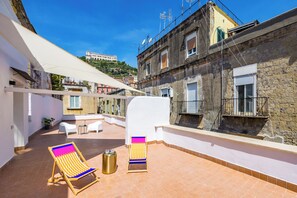 A rooftop terrace with two colorful sun loungers, a small table, and a trash can. The terrace is bordered by a white wall and a stone building.