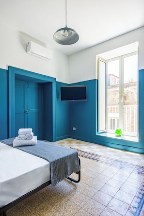 A hotel room with a bed, a TV mounted on the wall, a window with a view of the outside, and a blue door.