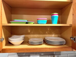 A wooden cabinet with two shelves. The top shelf holds colorful bowls and a blue cup. The bottom shelf contains stacked white bowls and plates. - Chicago 1BR Near Lake Michigan & Pratt Beach – Free Parking (Chicago)